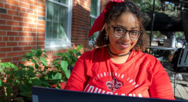 Courtney Johnson, wearing a red UL Lafayette shirt, sits outside at a coffee shop while working on her laptop.