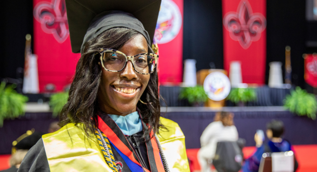 Denise Mallard, wearing her graduation cap and gown, smiles for a photo in front of the stage at Commencement.