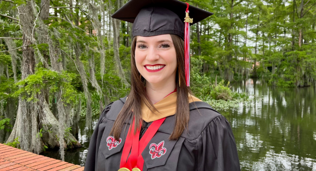 Kaley Carroll, wearing her graduation cap and gown, smiles for a photo in front of Cypress Lake on UL Lafayette's campus.