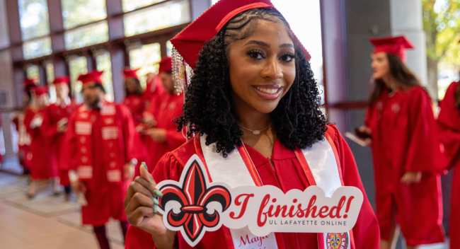Maya Major smiles at UL Lafayette Commencement ceremony, wearing a red cap and gown and holding a sign that reads: 'I finished!' 