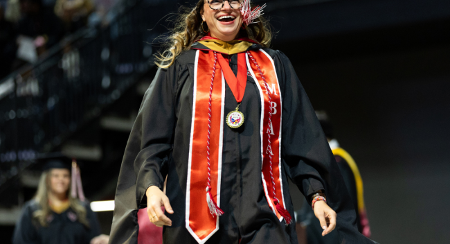 Kate Durio crosses the Commencement stage wearing a black cap and gown layered with a red MBA stole and tassels.