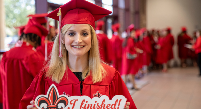 Online HPW graduate holds a UL Lafayette sign that reads 'I finished!' while smiling in her cap and gown.