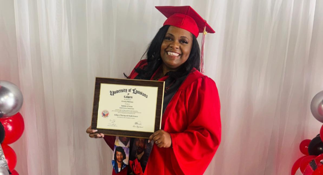 Health Services Administration online graduate smiles in cap and gown while holding her newly conferred bachelor's degree. 