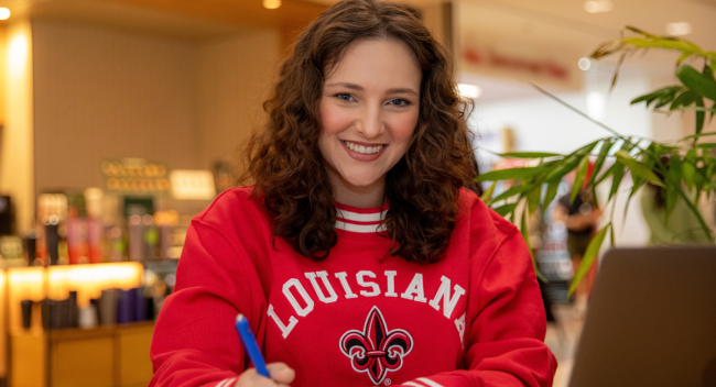 Brooke Perez, a student in the University of Louisiana at Lafayette's online criminal justice degree program, looks up at the camera and smiles as she works on her laptop in a coffee shop. 