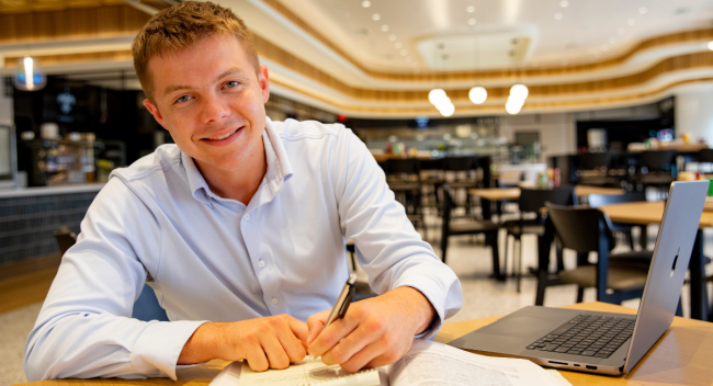 William Fralia, alumnus of UL Lafayette's sport management program, looks up at the camera and smile as he works on his laptop inside of a cafe.