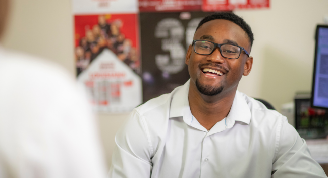 Andre Pittman, graduate of the University of Louisiana at Lafayette's sport management program, smiles as he speaks with a coworker at his desk.