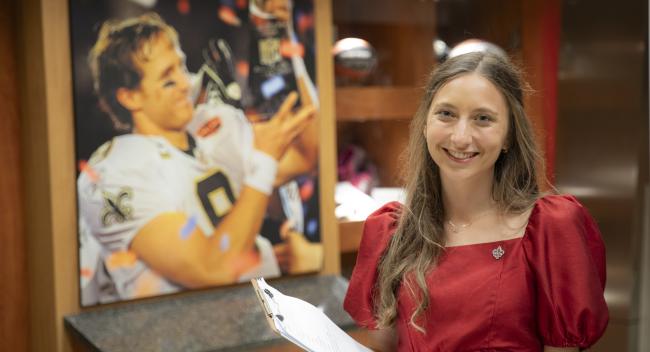 Sport Management program alumni Ashley Livaccari smiles holding a clipboard in front of a portrait of Drew Brees winning the Super Bowl.