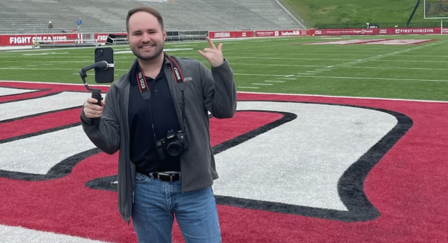 Daniel Richard, 2019 graduate of the University of Louisiana at Lafayette's marketing program, smiles for a picture on Cajun Field while capturing content with his phone.