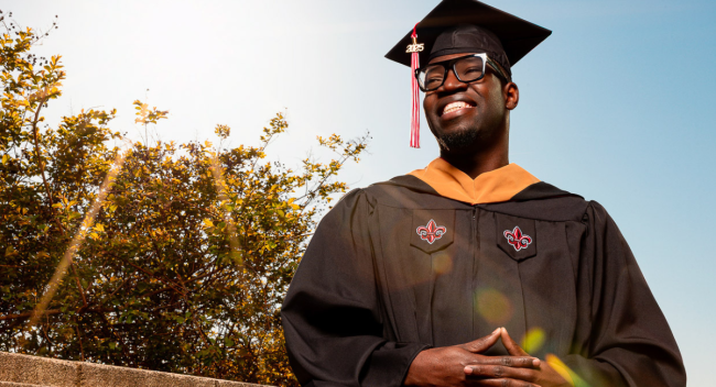 Mikenley Lundy, graduate of the University of Louisiana at Lafayette's online MBA program, smiles for a portrait in his master's regalia, the sun shining bright over his shoulder.