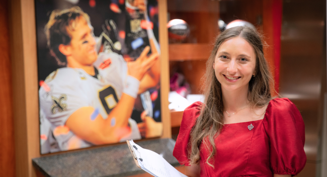 Sport Management program alumni Ashley Livaccari smiles holding a clipboard in front of a portrait of Drew Brees winning the Super Bowl.