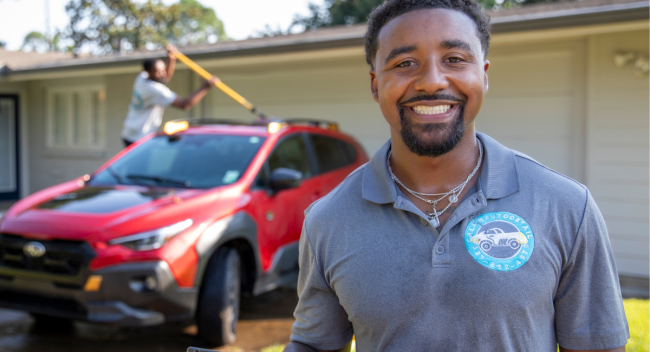 Entrepreneur Dillard Calais, graduate of UL Lafayette's online B.S.B.A. in Management program, smiles for a portrait while one of his employees details a client's car behind him.