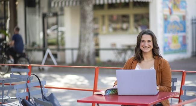 JoElle Judice is pictured outdoors at a red table with an open book and laptop. She has shoulder-length brown hair and is wearing a light brown blazer. 