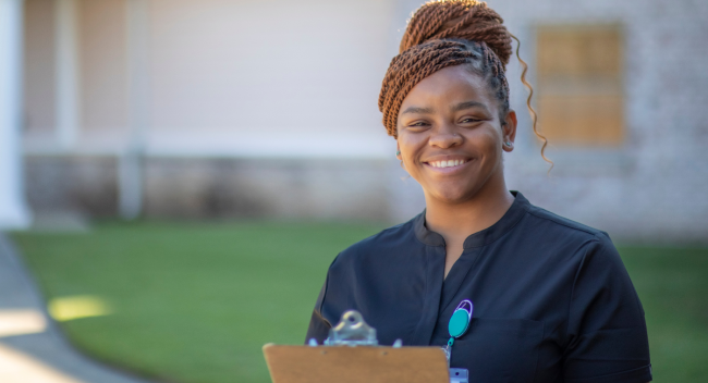 Destinee Francis, alumna of UL Lafayette's online bachelor's in health services administration program, smiles for a portrait in front of the facility she works in. She is wearing black scrubs and is holding a clipboard.
