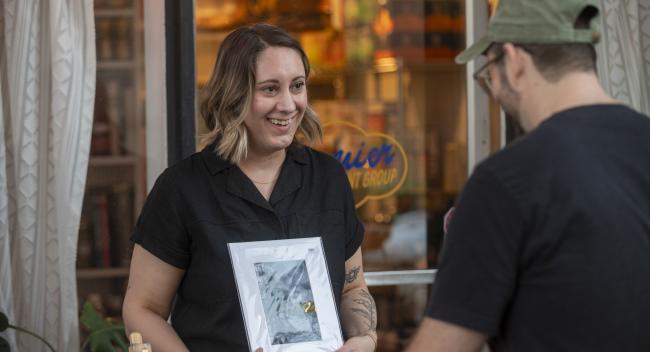 Lindsey Croall wears a black blouse and holds one of her watercolor illustrations while talking with a patron during an ArtWalk event.