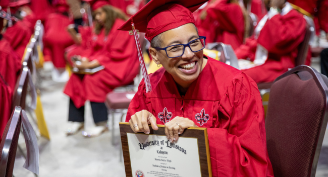 Online RN to BSN graduate smiles in cap and gown holding her newly earned degree.