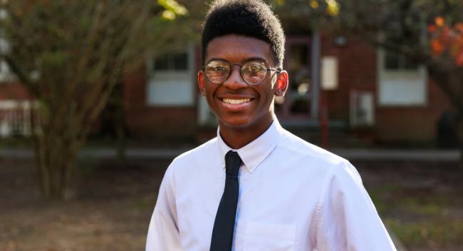 Portrait photograph of Xzayvion Gaye smiling wearing a white long sleeve button down dress shirt and a black tie. He also is wearing glasses standing in front of trees and a building.
