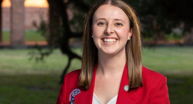 Portrait photograph of Mary McMahon smiling in front a campus building. She has on a red blazer with a University logo and a white blouse.