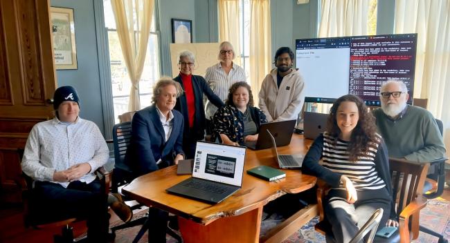 A group of eight people sit at a table in a conference room with a laptop and TV screen behind them.