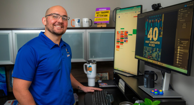 Grayson Stepanek is pictured wearing a blue polo shirt and smiling at his office desk at Goodwill Acadiana.