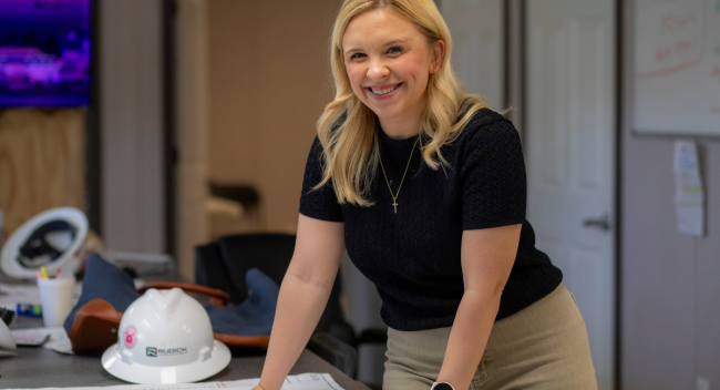 Jillian Blanchard smiles into the camera while leaning over a table laden with construction plans and a Rudick Construction Group hard hat.