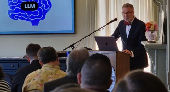 Man in suit and bowtie stands behind a lecturn with a screen behind him featuring an illustration of a brain and the letters LLM.