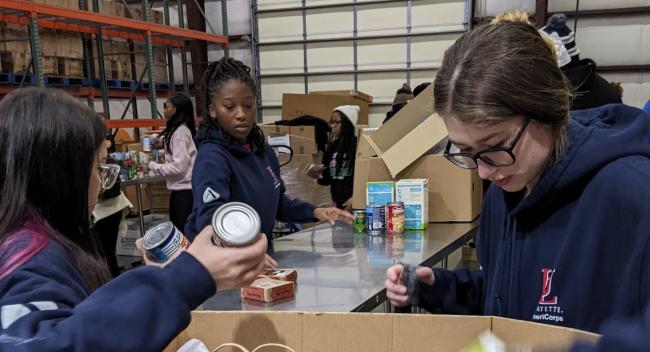 Three students in blue hoodies sort canned goods at a Second Harvest warehouse.