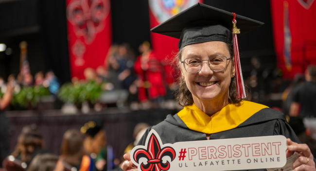 Karen LeDoux is pictured during commencement ceremonies wearing UL Lafayette's black master's cap and gown and holding a sign that reads: #PERSISTENT UL Lafayette Online.