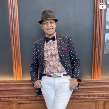 An older man in a polka dot jacket, white pants, gucci belt, floral shirt, black bowtie, red pocketsquare, and a fedora leans casually against a wood paneled wall. 