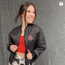 A young woman with brown hair smiles while looking off to the side, she wears a hip outfit centered on a quilted black jacket with a fleur-de-lis on the breast.