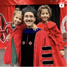 A UL Lafayette doctoral graduate stands with his small daughters, they all wear red. Icon in corning indicates video.