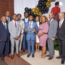Cross section of formally dressed men and women at the inaugural Black History Luncheon. Young guy in the middle is holding up an award plaque 