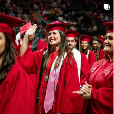 A white female graduate waves happily to someone in the stands at Commencement. She wears a red cap and gown and pink stole.
