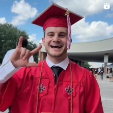 A white male UL Lafayette graduate makes a UL hand sign with a wide smile. He wears a red cap and gown.