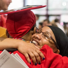A Black female wearing glasses tightly hugs a UL Lafayette graduate in red cap and gown.