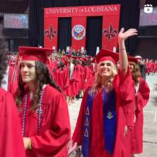 Lines of commencing UL Lafayette grads can be seen in red caps and gowns. A white female waves enthusiastically to someone behind and above the photographer.
