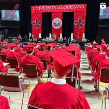 Rows of seated graduates in red caps and gowns can be seen from behind with the University of Louisiana Commencement stage seen in the distance.