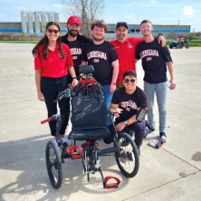 A group of students in Louisiana Ragin' Cajuns shirts gather around a three-wheeled motorized vehicle.