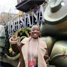 A woman smiling and holding up a UL Handsign while standing next to a  Mardis-Gras float. 