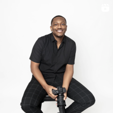 A black man sitting holding a camera infront of a white background