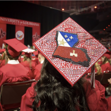 A student wearing red graduation robes with a focus on their detailed hat with the outline of louisiana on it