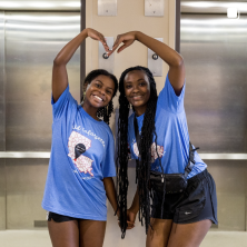 Two women in matching blue shirts posing together to make a giant heart with their hands