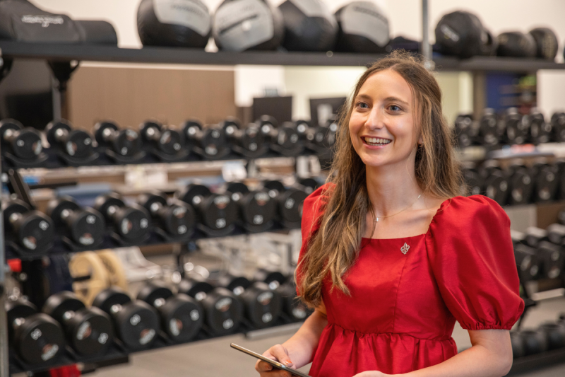 Ashley Livaccari, graduate of the sport management degree program at UL Lafayette, smiles while holding clipboard in the New Orleans Pelicans weight room.