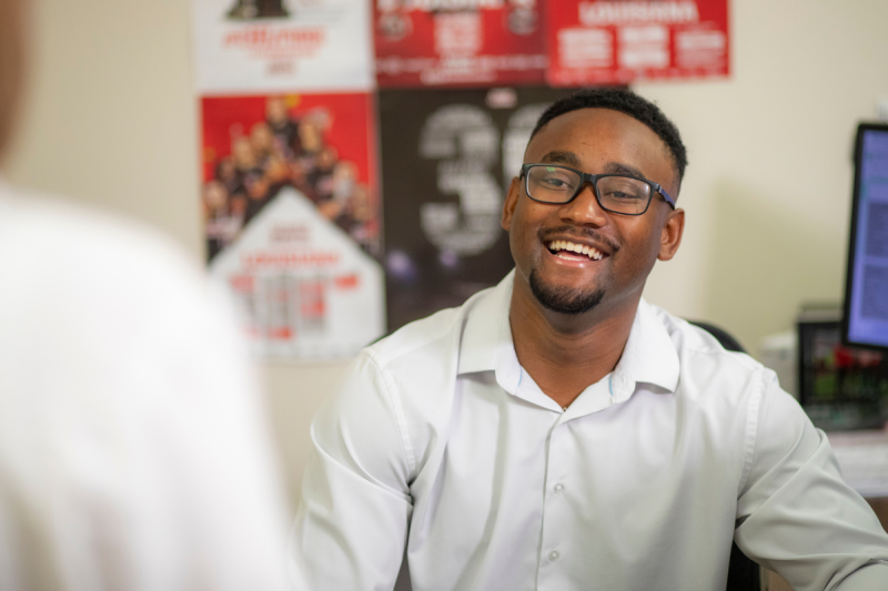 Andre Pittman, graduate of the University of Louisiana at Lafayette's sport management program, smiles as he speaks with a coworker at his desk.