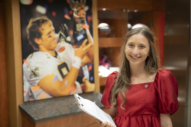 Sport Management program alumni Ashley Livaccari smiles holding a clipboard in front of a portrait of Drew Brees winning the Super Bowl.