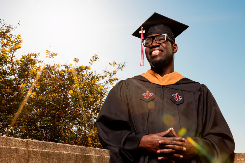 Mikenley Lundy, graduate of the University of Louisiana at Lafayette's online MBA program, smiles for a portrait in his master's regalia, the sun shining bright over his shoulder.