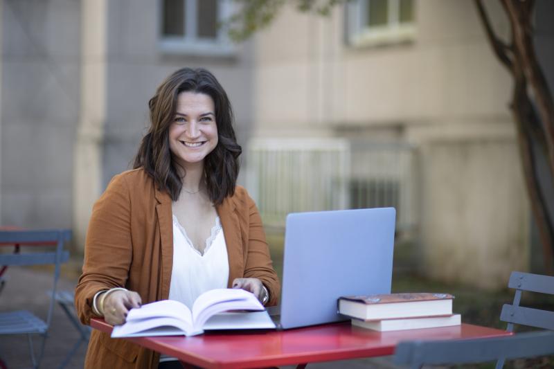 JoElle Judice is pictured outdoors at a red table with an open book, laptop, and stack of books. She has shoulder-length brown hair and is wearing a light brown blazer. 