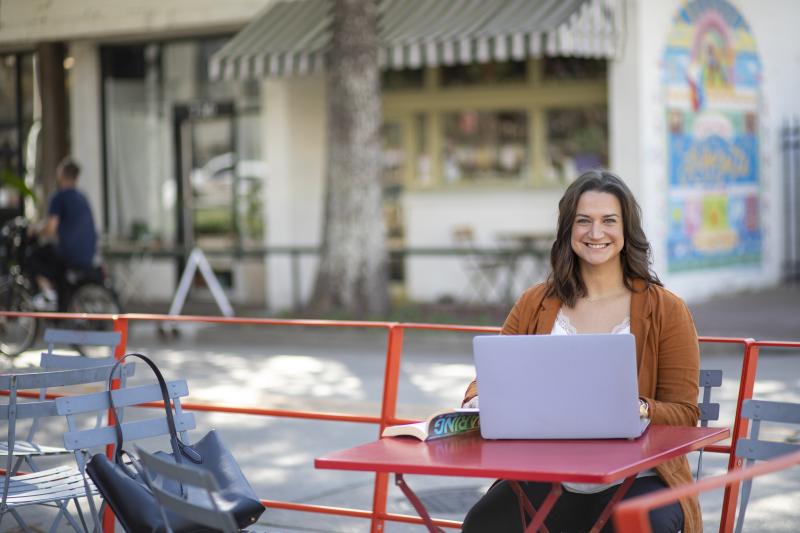 JoElle Judice is pictured outdoors at a red table with an open book and laptop. She has shoulder-length brown hair and is wearing a light brown blazer. 