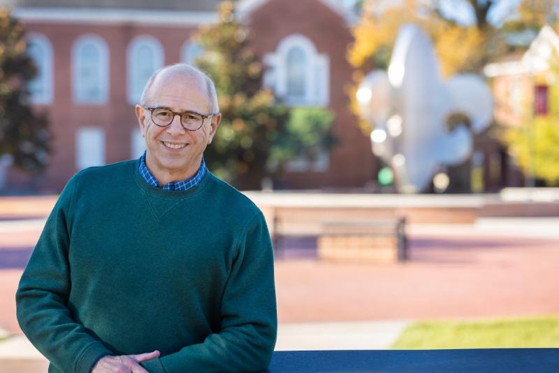 A man wearing glasses and a green sweater smiles in front of the fleur de lis fountain in the UL Lafayette quad.