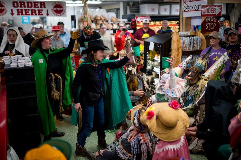 Woman in a cowboy hat and green cape leads a crowd of costumed Mardi Gras revelers in a chant.