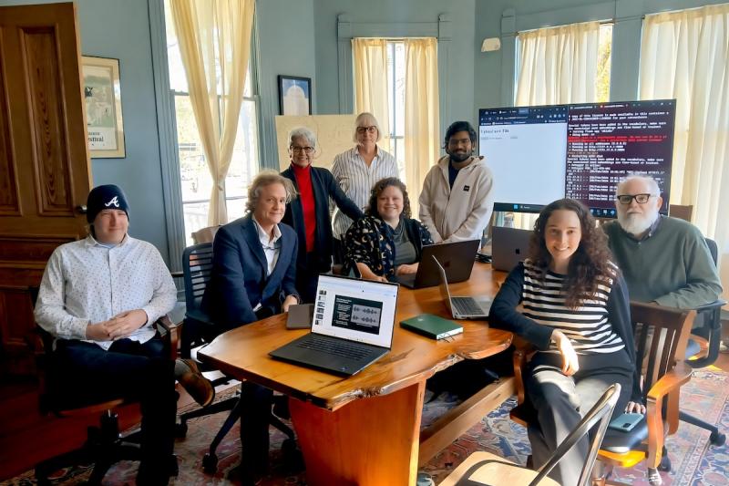 A group of eight people sit at a table in a conference room with a laptop and TV screen behind them.